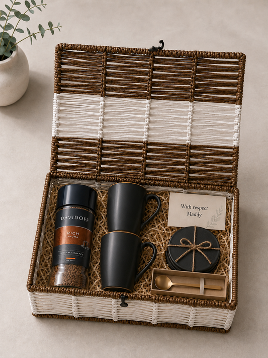 A reusable white and brown trunk basket featuring Davidoff Rich Aroma coffee, two matte black mugs, black coasters, a gold spoon, a message card, and shredded paper packaging.