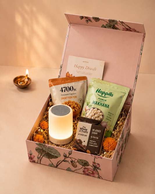 Diwali Cheer Hamper with a portable Bluetooth speaker, sweet and salty popcorn, Himalayan salt and pepper makhana, cocoa protein bar, and Diwali greeting card in a printed gift box.