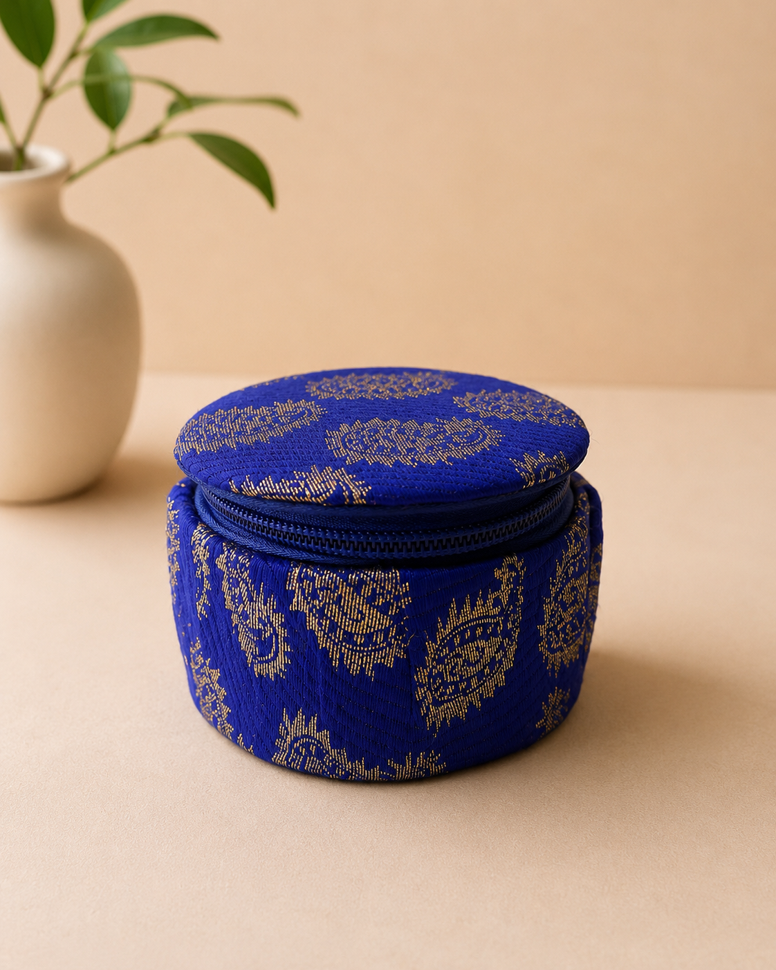 A small round blue fabric dabbi with traditional gold keri print and a zip closure, suitable for jewellery, chocolates, dry fruits, and festive gifting.