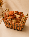 A rustic woven basket featuring a Lumba Rakhi, scented candle, fragrance mist, bath and body products, crochet soft toy, fabric gift, and floral styling for Bhabhi Raksha Bandhan gifting.