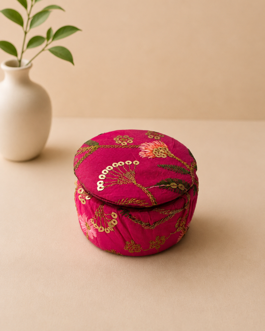 A small round fuchsia fabric dabbi with gold sequin embroidery and a fitted lid, ideal for presenting jewellery, chocolates, dry fruits, and festive keepsakes.