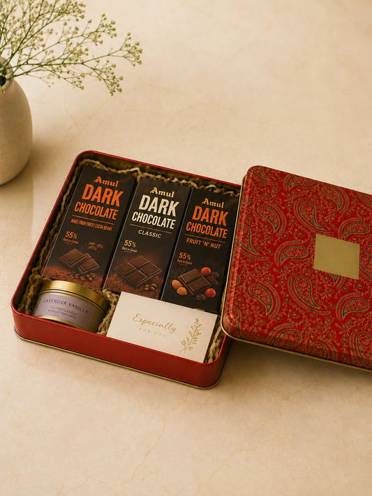 A red printed tin gift box featuring Amul dark chocolates, a lavender vanilla scented candle, a note card, and shredded paper packaging for festive and thoughtful gifting.