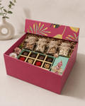 A pink floral designer gift box with assorted dry fruits, a luxury chocolate box, a custom note card, and shredded paper packaging for festive and thoughtful gifting.